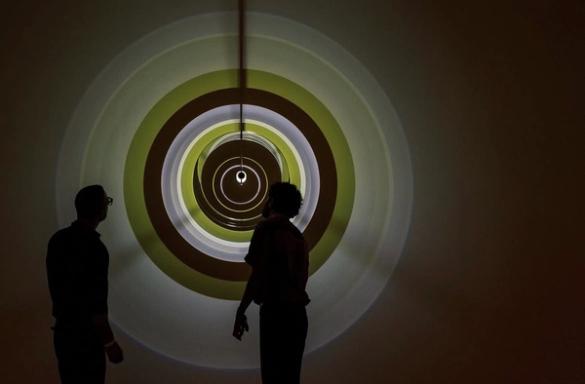 Two silhouetted people look at a wall projection of concentric green, yellow, and brown circles forming a deep tunnel.