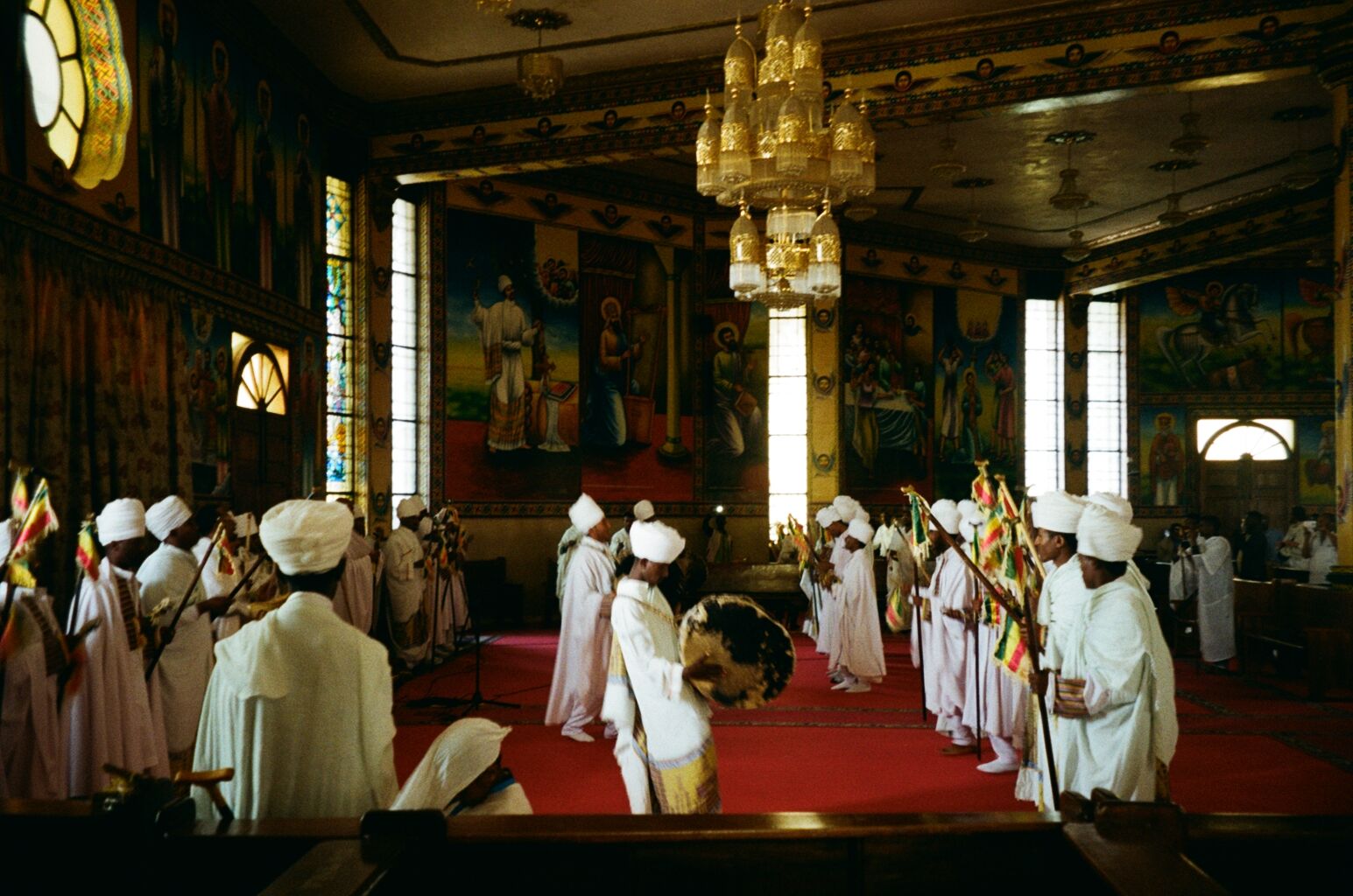 People in white robes perform a ceremony with flags and a drum inside a church decorated with vibrant murals and a golden chandelier.