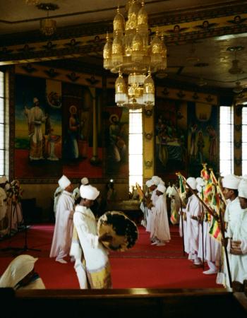 People in white robes perform a ceremony with flags and a drum inside a church decorated with vibrant murals and a golden chandelier.