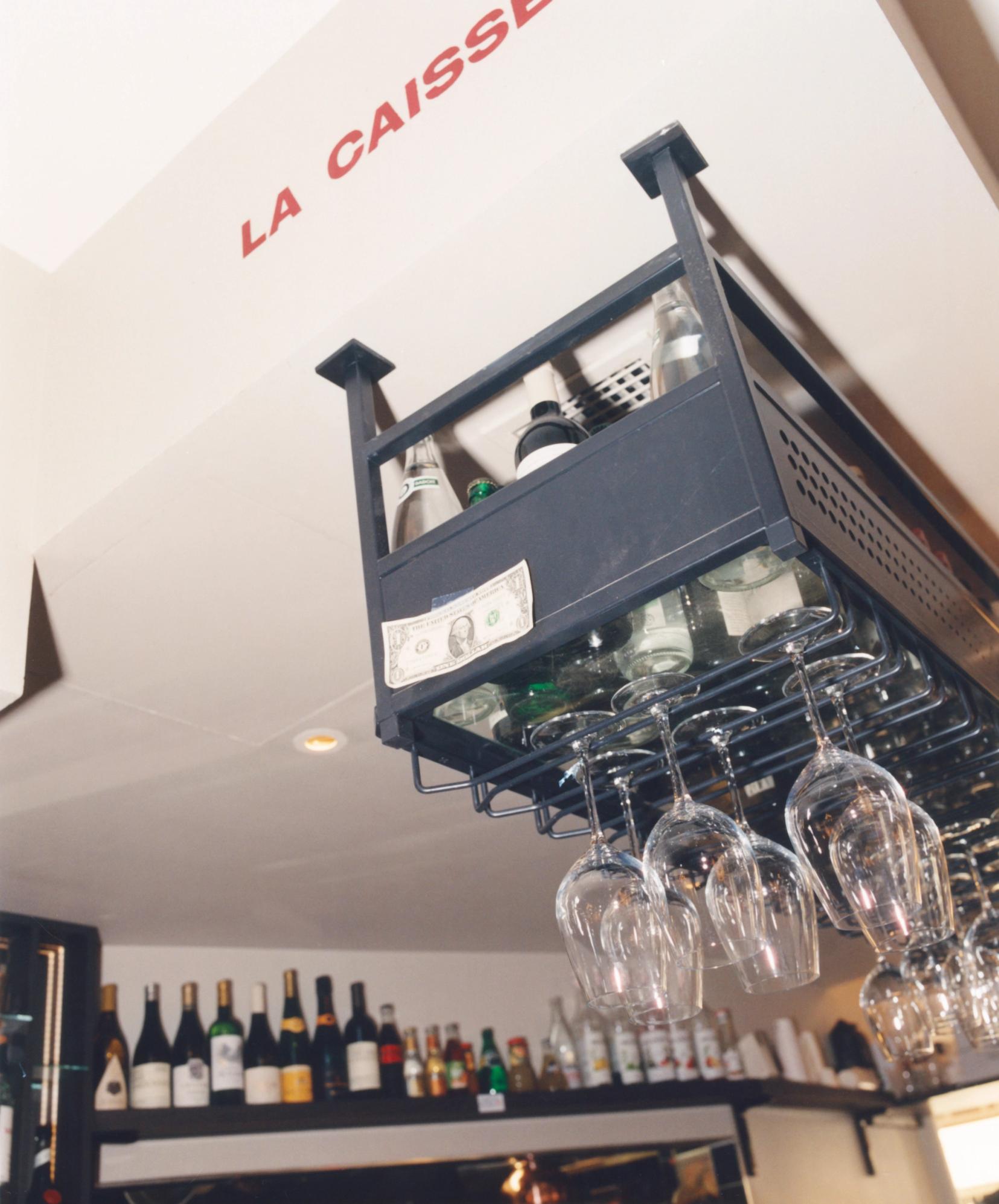 Black overhead bar rack with bottles, hanging wine glasses, and a tucked-in dollar bill, beneath a ceiling reading 'LA CAISSE' in red.