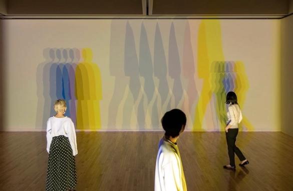 Three people view a projection art installation featuring layered, colorful silhouettes of figures on a wall.