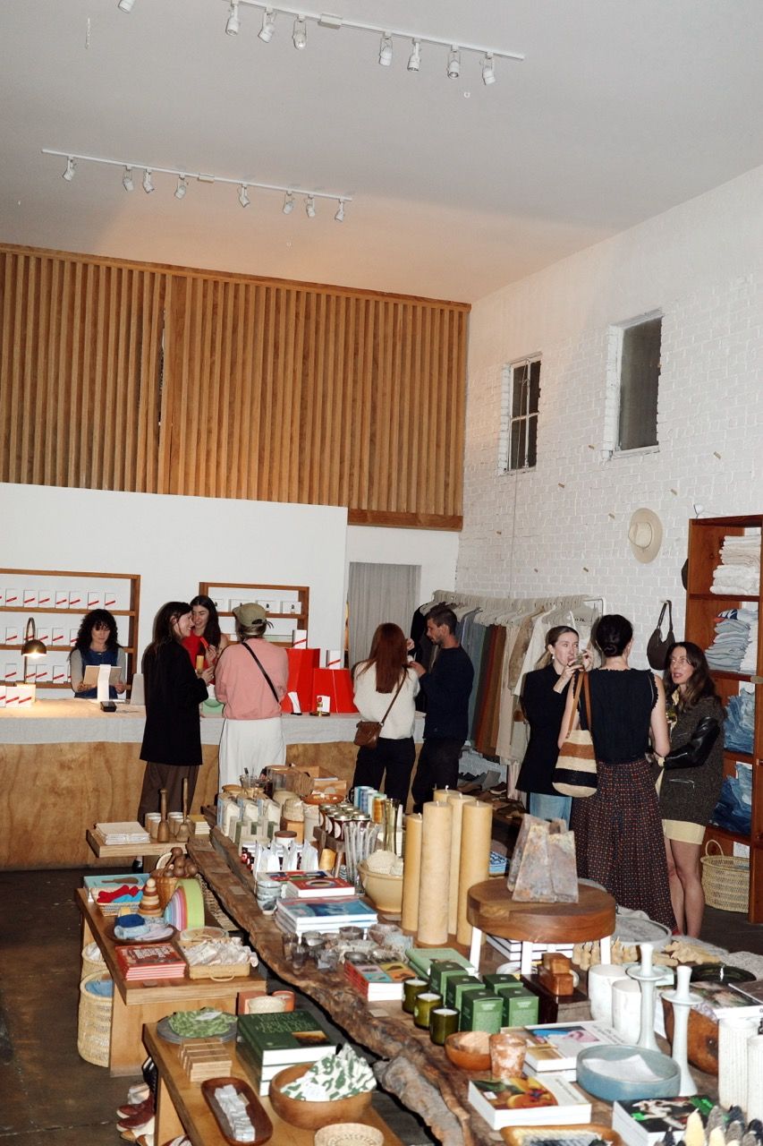 Inside a bustling modern boutique with white walls and wood accents, people browse clothing racks and rustic tables filled with candles, books, and home goods.