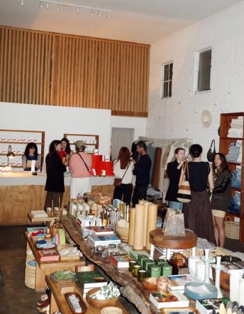 Inside a bustling modern boutique with white walls and wood accents, people browse clothing racks and rustic tables filled with candles, books, and home goods.