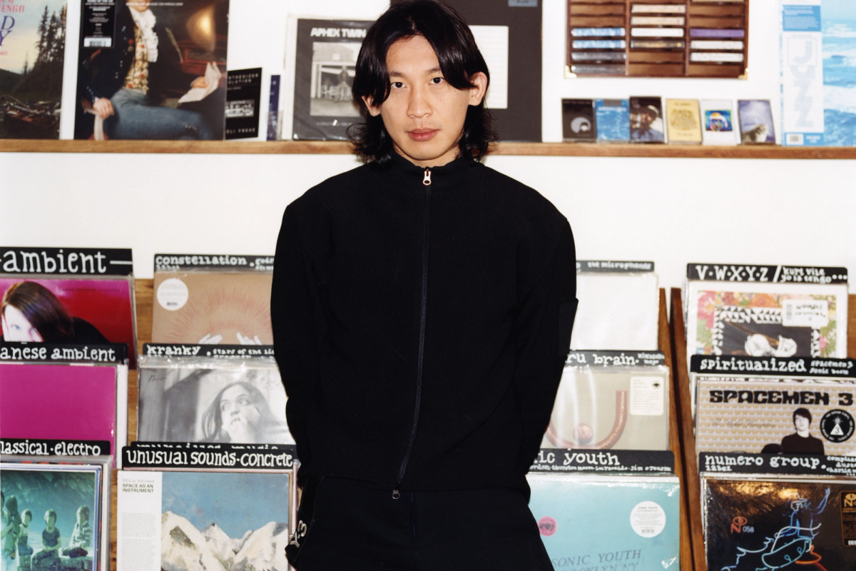 An Asian man with long dark hair stands in a record store in front of shelves filled with vinyl albums.