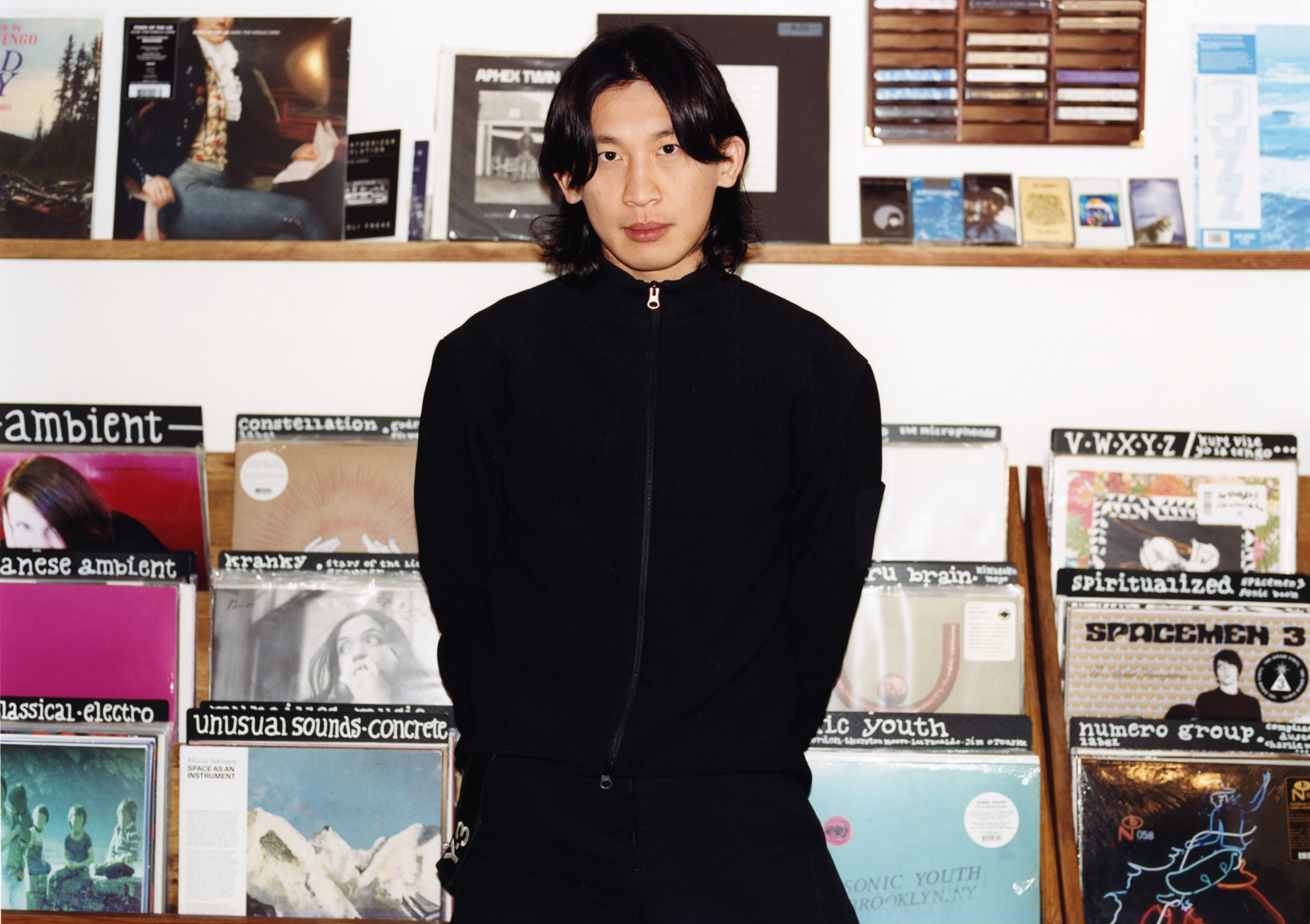 A man with long dark hair and a black jacket stands in front of shelves of vinyl records.