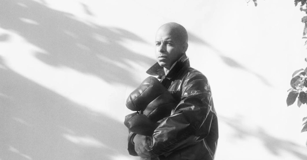 Black and white image of a person with a shaved head wearing a dark jacket, holding a bundled item, standing against a sunlit wall with leaf shadows.