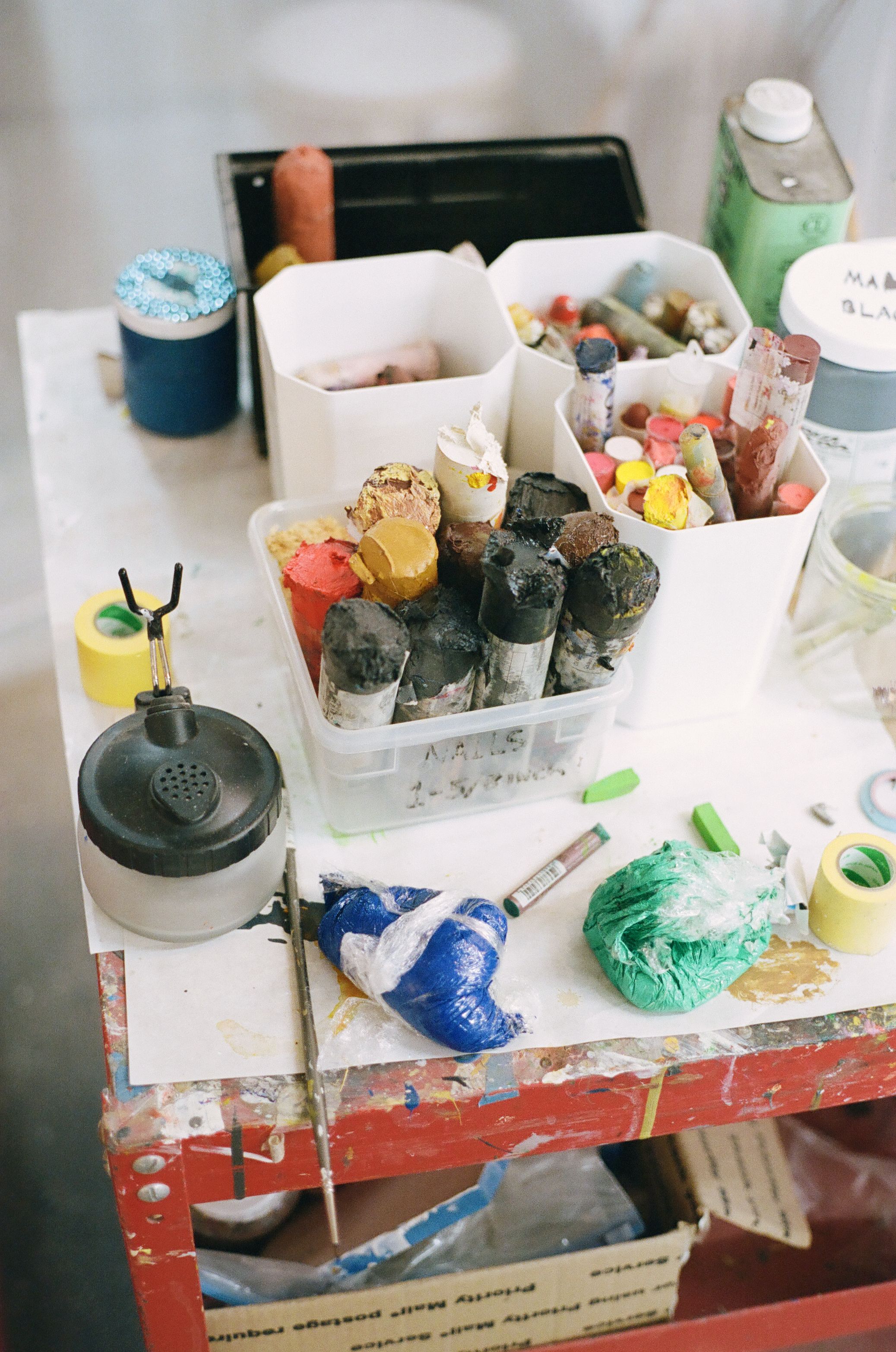 A busy artist's table covered with containers of colorful paints, pastels, and assorted art supplies.