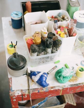 A busy artist's table covered with containers of colorful paints, pastels, and assorted art supplies.