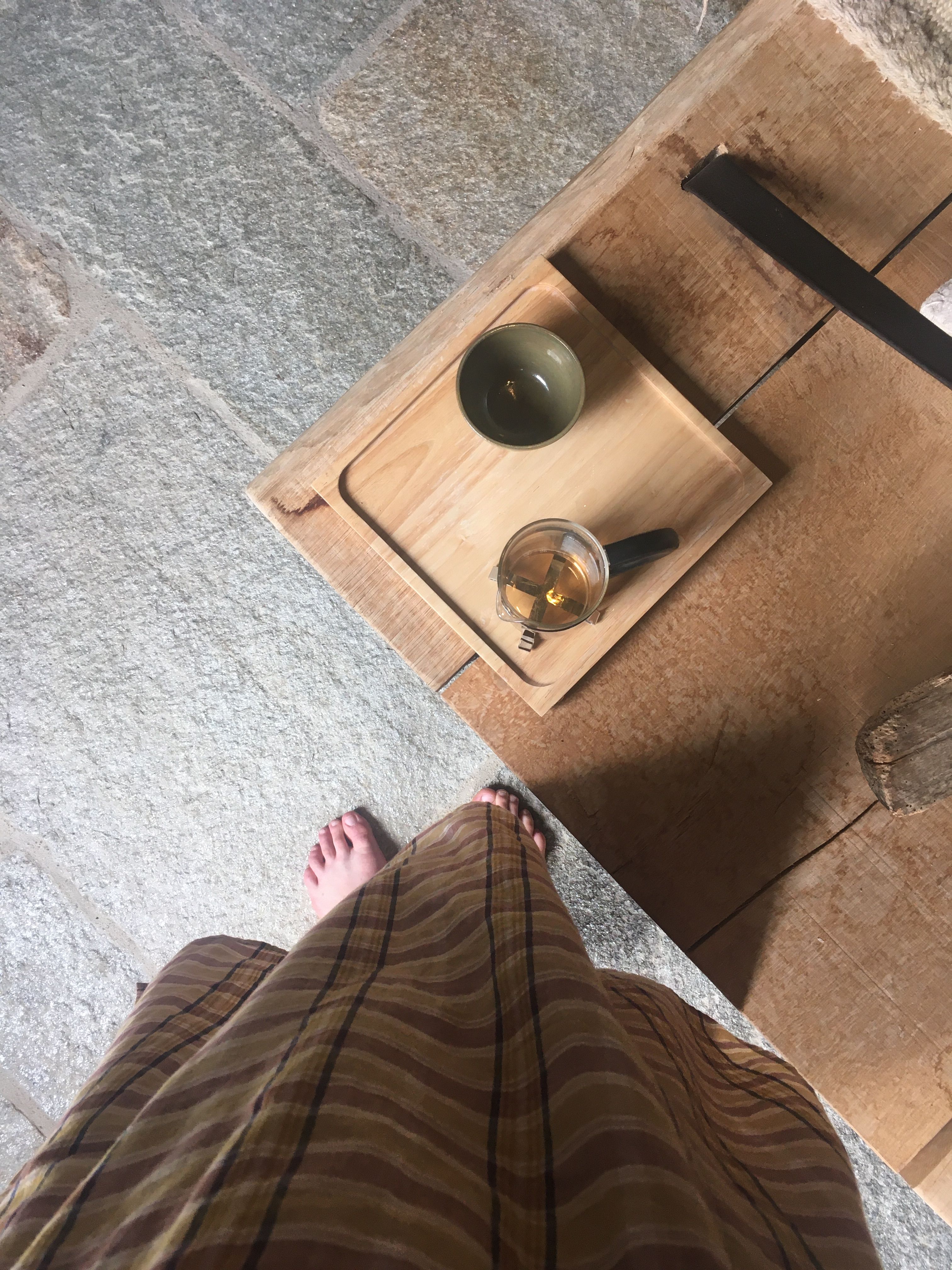 Overhead view of bare feet and a brown striped skirt on a stone floor, beside a wooden table with a tea tray holding a green cup and glass teapot.