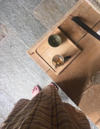 Overhead view of bare feet and a brown striped skirt on a stone floor, beside a wooden table with a tea tray holding a green cup and glass teapot.