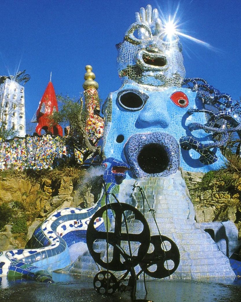 A fantastical outdoor sculpture garden with a large blue mosaic face fountain, a mirrored head sculpture, and a kinetic metal artwork.