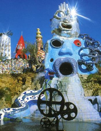 A fantastical outdoor sculpture garden with a large blue mosaic face fountain, a mirrored head sculpture, and a kinetic metal artwork.