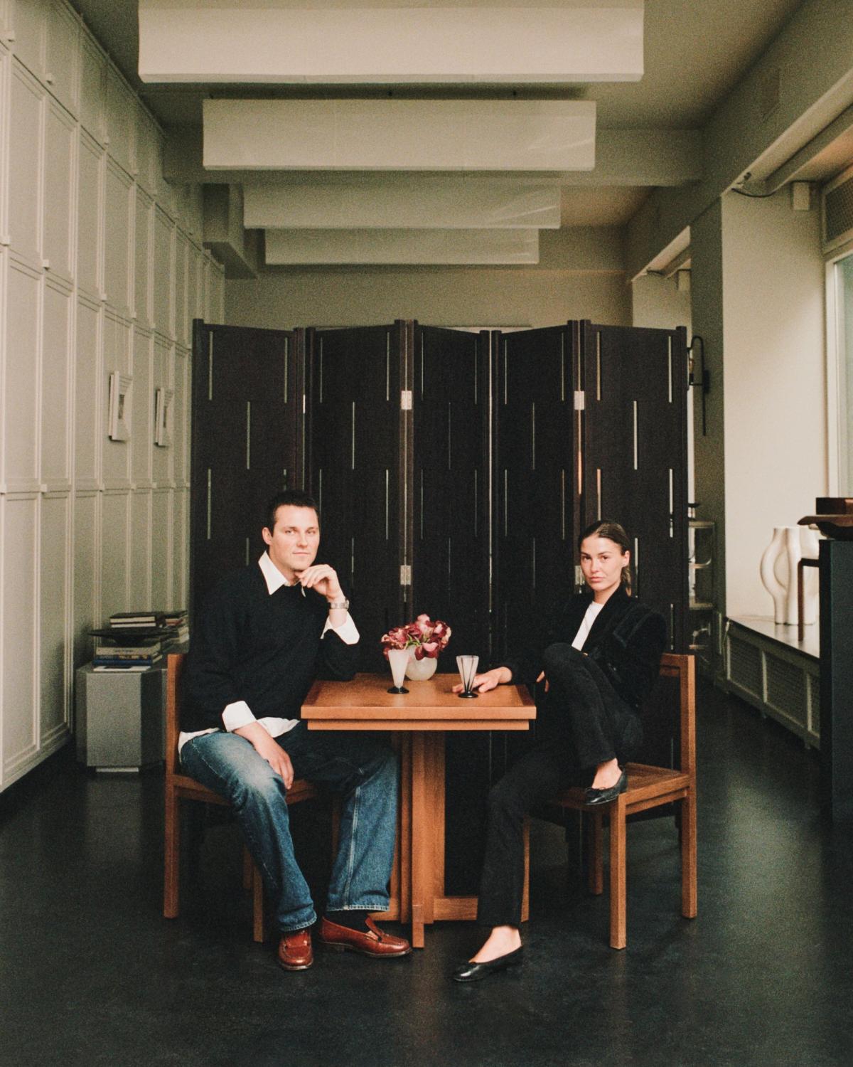 A man and a woman sit at a small wooden table, facing the camera. He wears a dark sweater and jeans, she wears a dark blazer and pants. A dark room divider stands behind them.