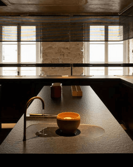 Dark, minimalist counter with a curved bronze faucet flowing water into an orange bowl in a shallow basin, against windows with metal blinds.