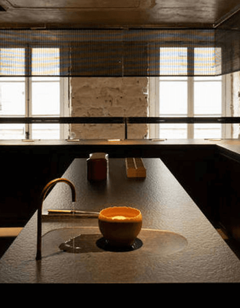 Dark, minimalist counter with a curved bronze faucet flowing water into an orange bowl in a shallow basin, against windows with metal blinds.