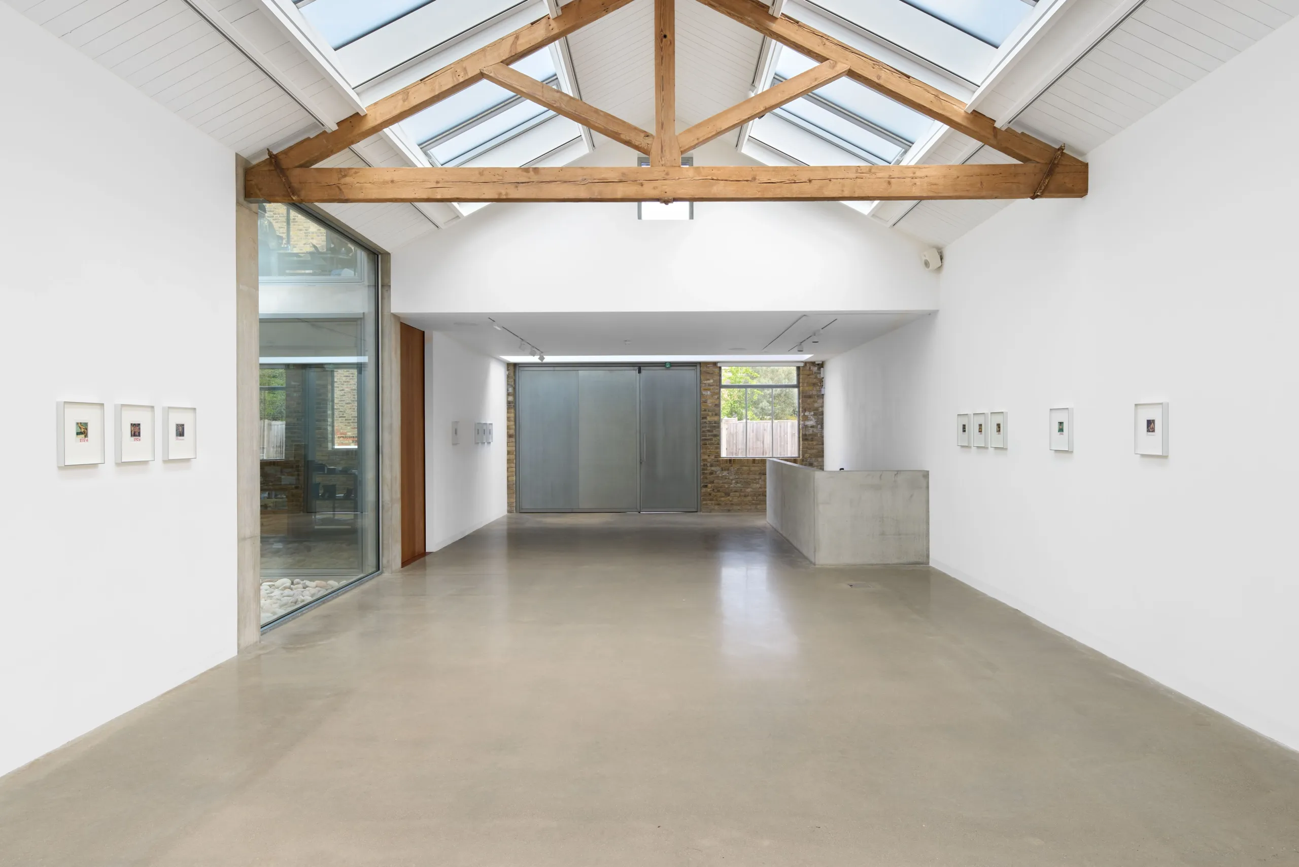 Modern art gallery with high vaulted ceiling, exposed wooden beams, skylights, concrete floor, and small framed artworks on white walls.