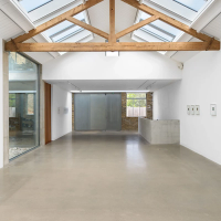 Modern art gallery with high vaulted ceiling, exposed wooden beams, skylights, concrete floor, and small framed artworks on white walls.