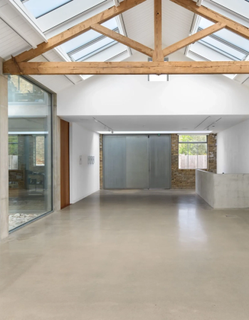 Modern art gallery with high vaulted ceiling, exposed wooden beams, skylights, concrete floor, and small framed artworks on white walls.