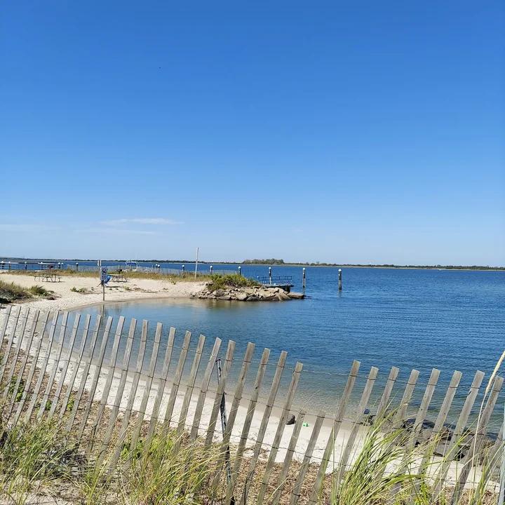 A sandy beach with a wooden fence in the foreground, calm bay water, and a small dock under a clear blue sky.