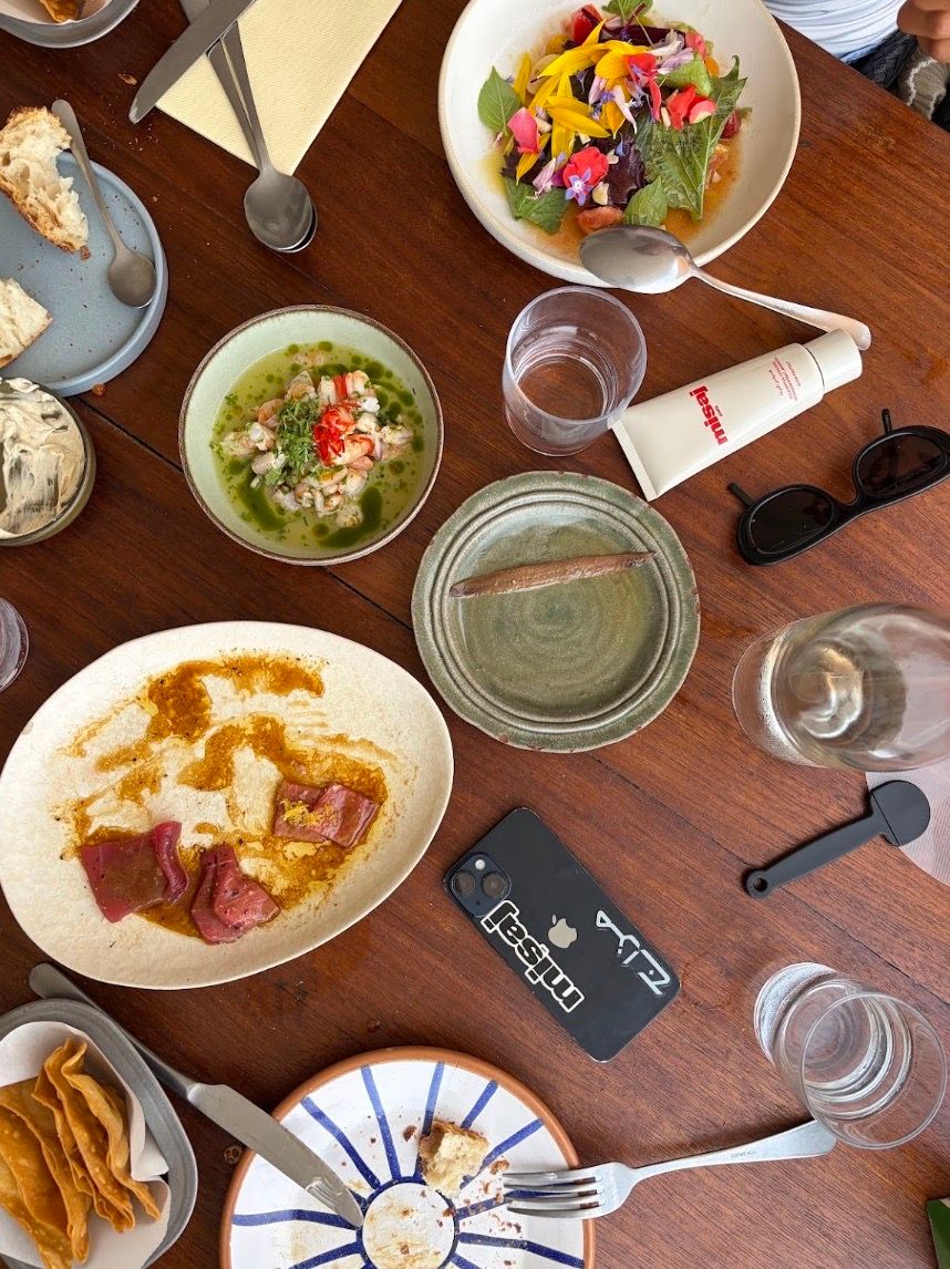 Overhead view of a wooden dining table filled with a colorful floral salad, ceviche, partially eaten dishes, glasses, cutlery, a phone, and sunglasses.