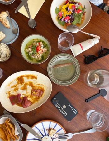 Overhead view of a wooden dining table filled with a colorful floral salad, ceviche, partially eaten dishes, glasses, cutlery, a phone, and sunglasses.