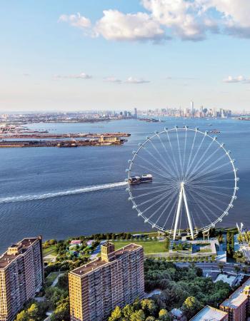 Aerial view of a large Ferris wheel on a waterfront with a baseball field, residential buildings, and the Manhattan skyline in the distance.