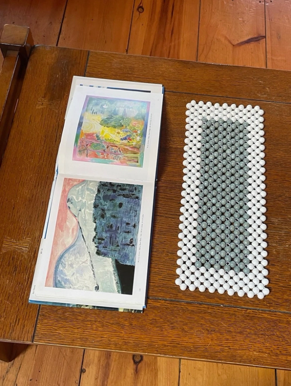 An open art book showing two colorful paintings and a white and gray beaded mat on a wooden table.