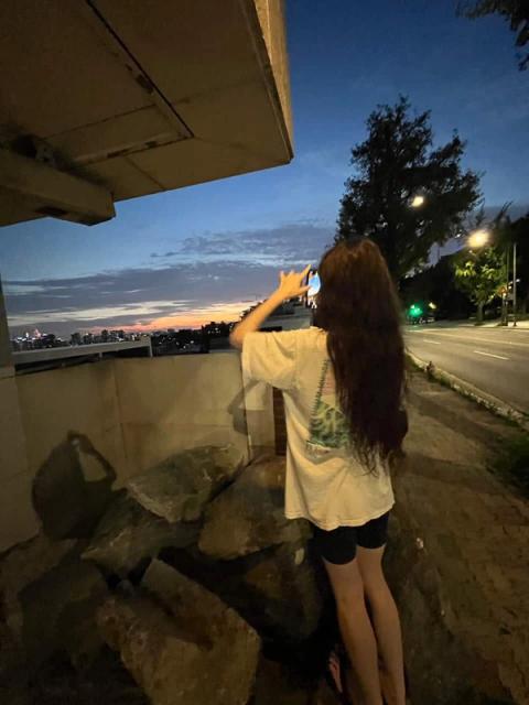 A person with long dark hair photographs a colorful sunset over a distant city from under a concrete overpass.