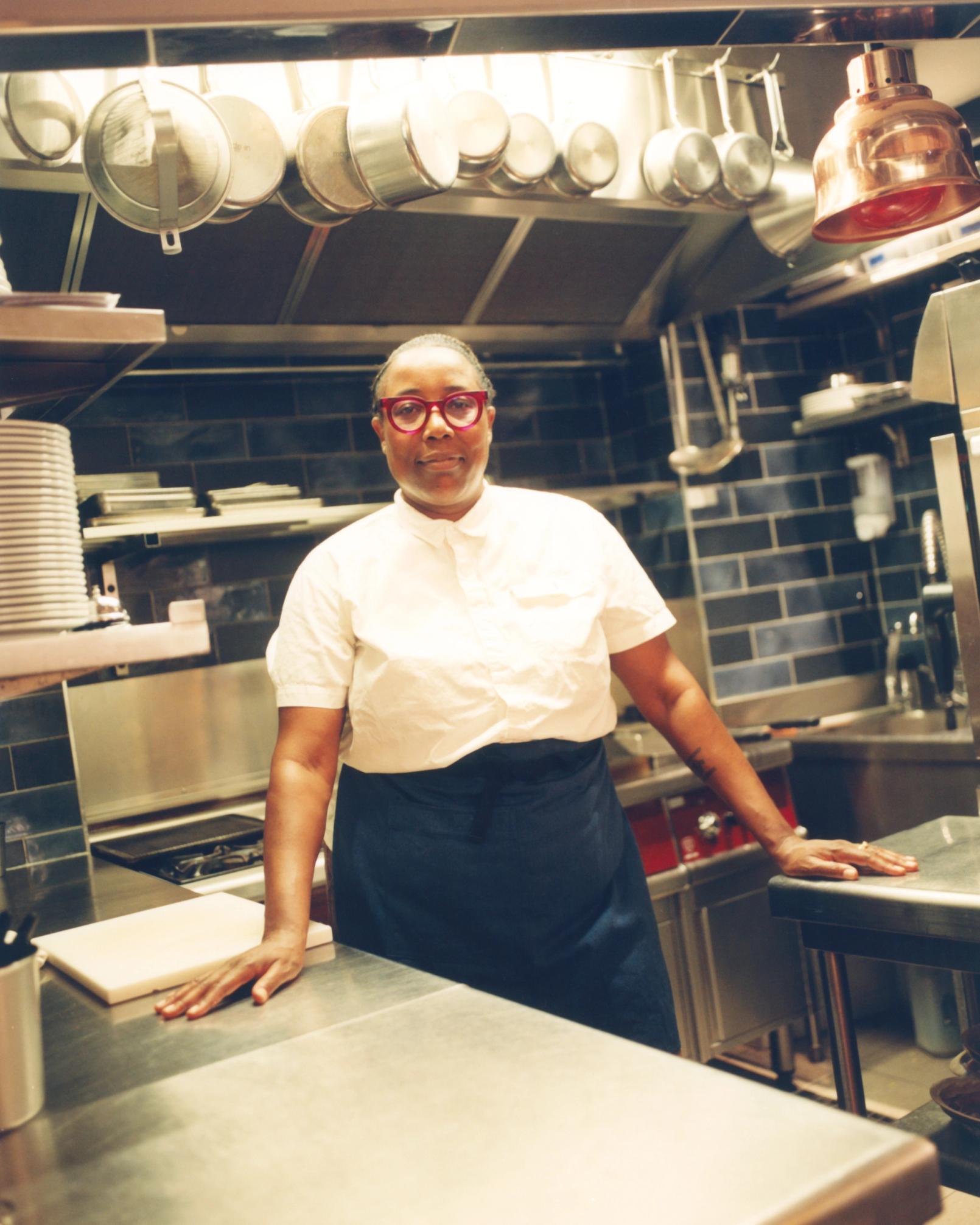 A Black woman in red glasses, a white shirt, and a dark apron stands in a commercial kitchen, hands resting on a stainless steel counter.