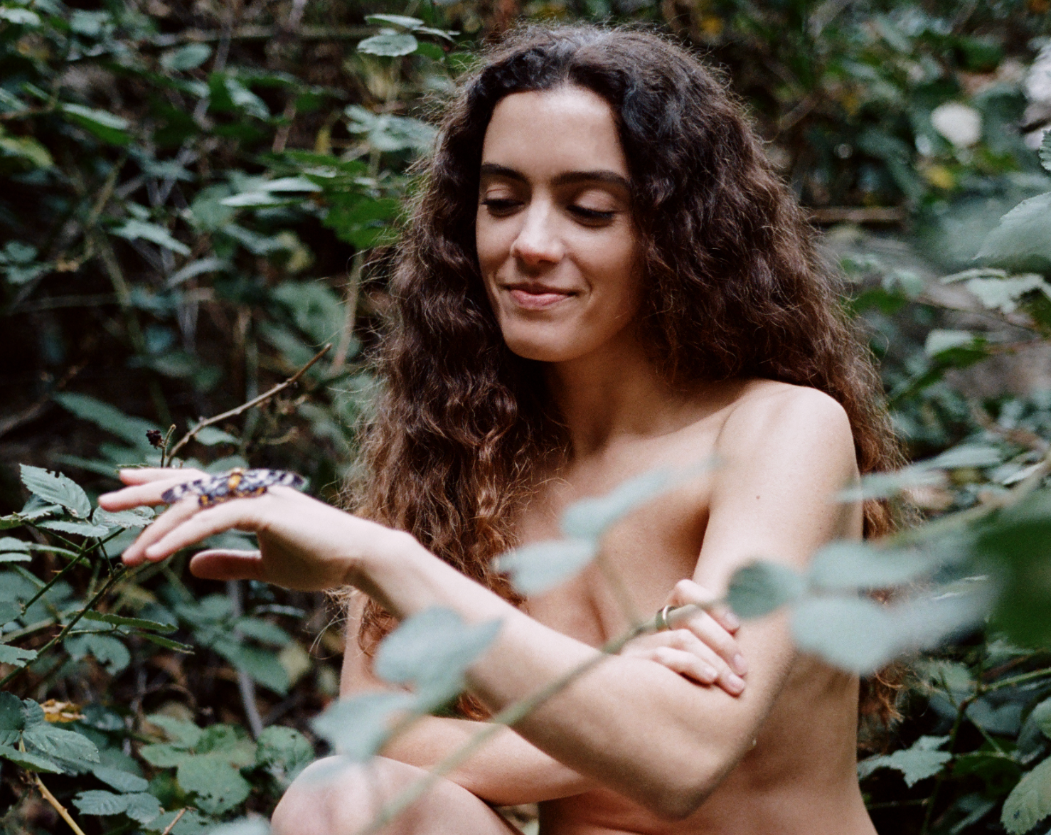 A bare-chested woman with long curly hair smiles at a patterned lizard on her hand, surrounded by green foliage.