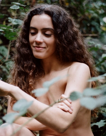 A bare-chested woman with long curly hair smiles at a patterned lizard on her hand, surrounded by green foliage.