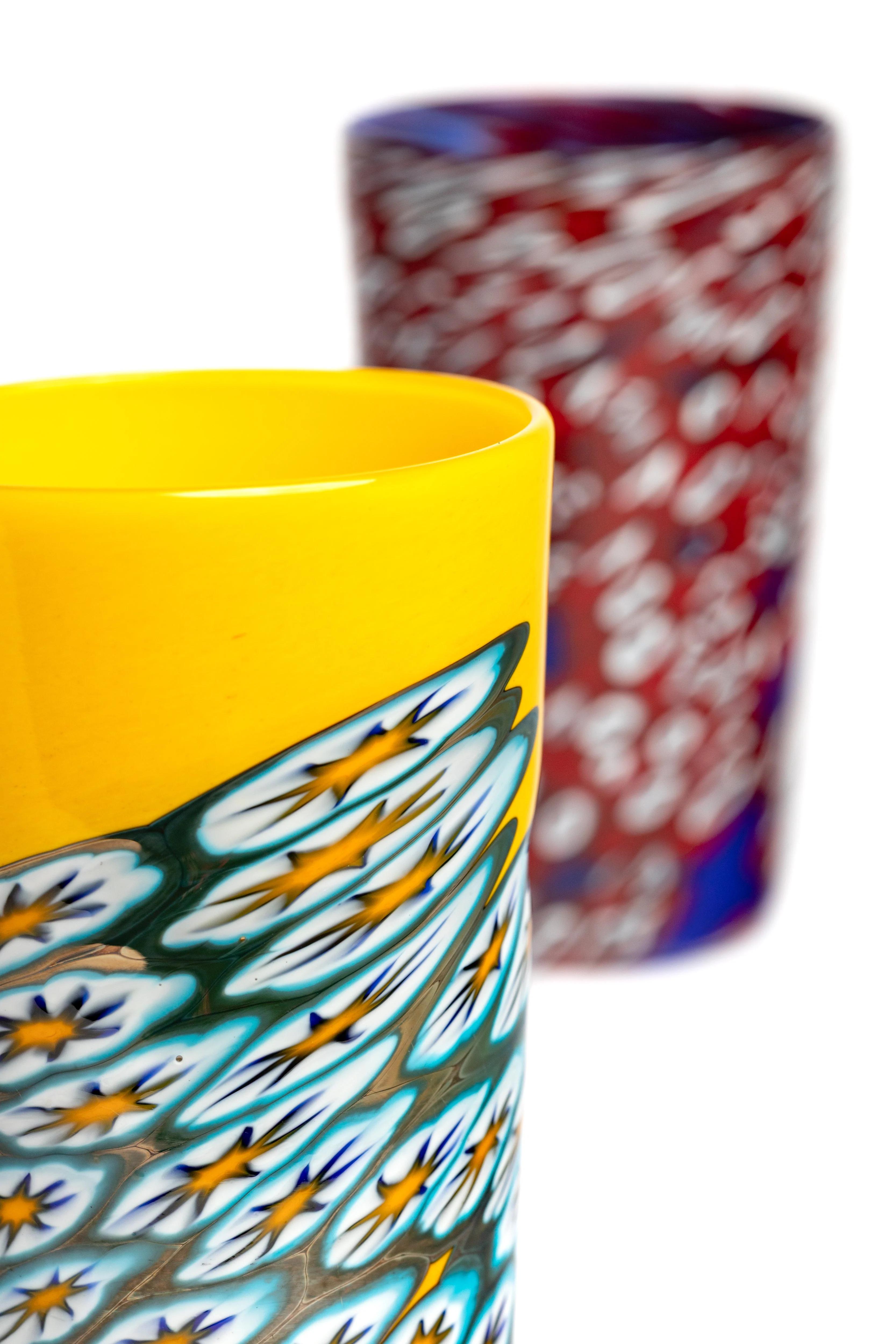 A yellow glass tumbler with a detailed white, blue, and yellow pattern, partially in front of a blurry red and white flecked glass.