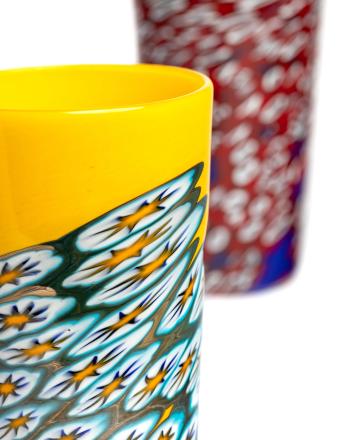 A yellow glass tumbler with a detailed white, blue, and yellow pattern, partially in front of a blurry red and white flecked glass.