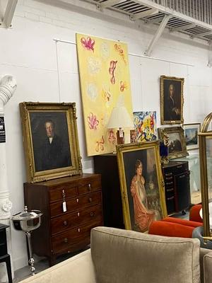 Numerous framed paintings, including portraits, landscapes, and an abstract yellow piece, are displayed on a white wall alongside a wooden chest of drawers and a beige sofa.