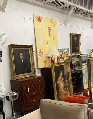 Numerous framed paintings, including portraits, landscapes, and an abstract yellow piece, are displayed on a white wall alongside a wooden chest of drawers and a beige sofa.