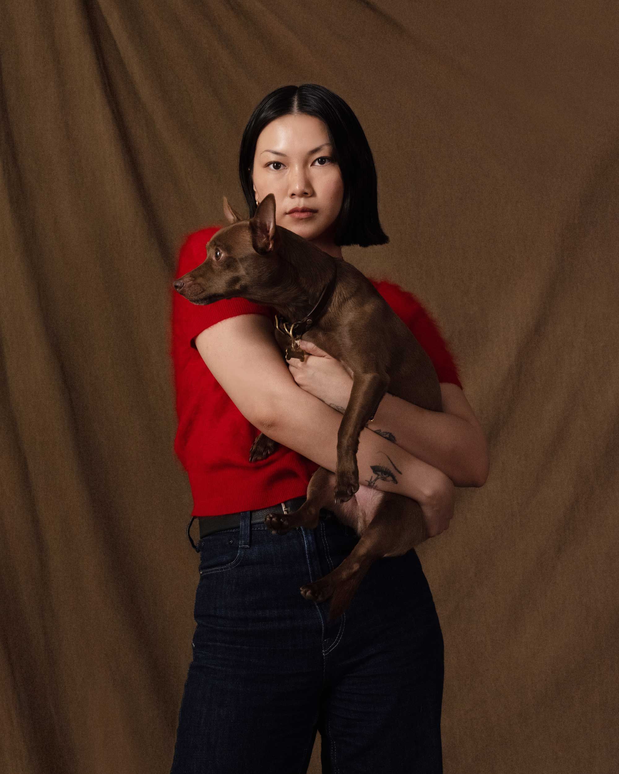An East Asian woman in a red top holds a small brown dog against a brown background.