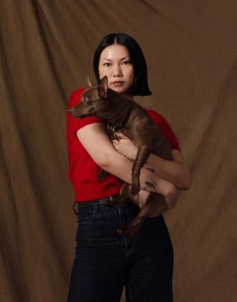 An East Asian woman in a red top holds a small brown dog against a brown background.