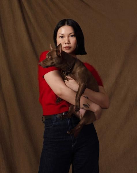A woman with short dark hair and a red top holds a small brown dog against a brown background.