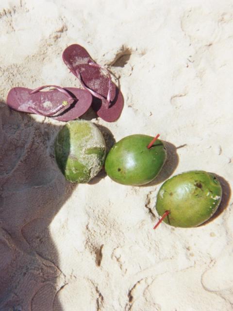 Flip-flops and three green coconuts, two with straws, on a sandy beach.