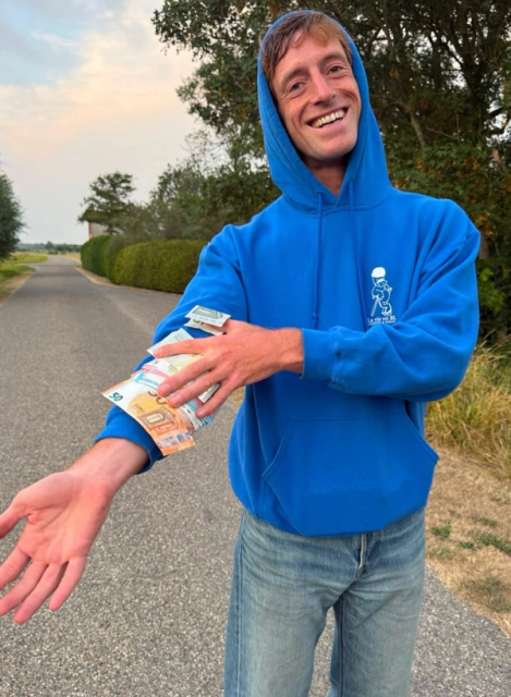 A smiling man in a blue hoodie displays Euro banknotes on his arm while extending his other hand forward.