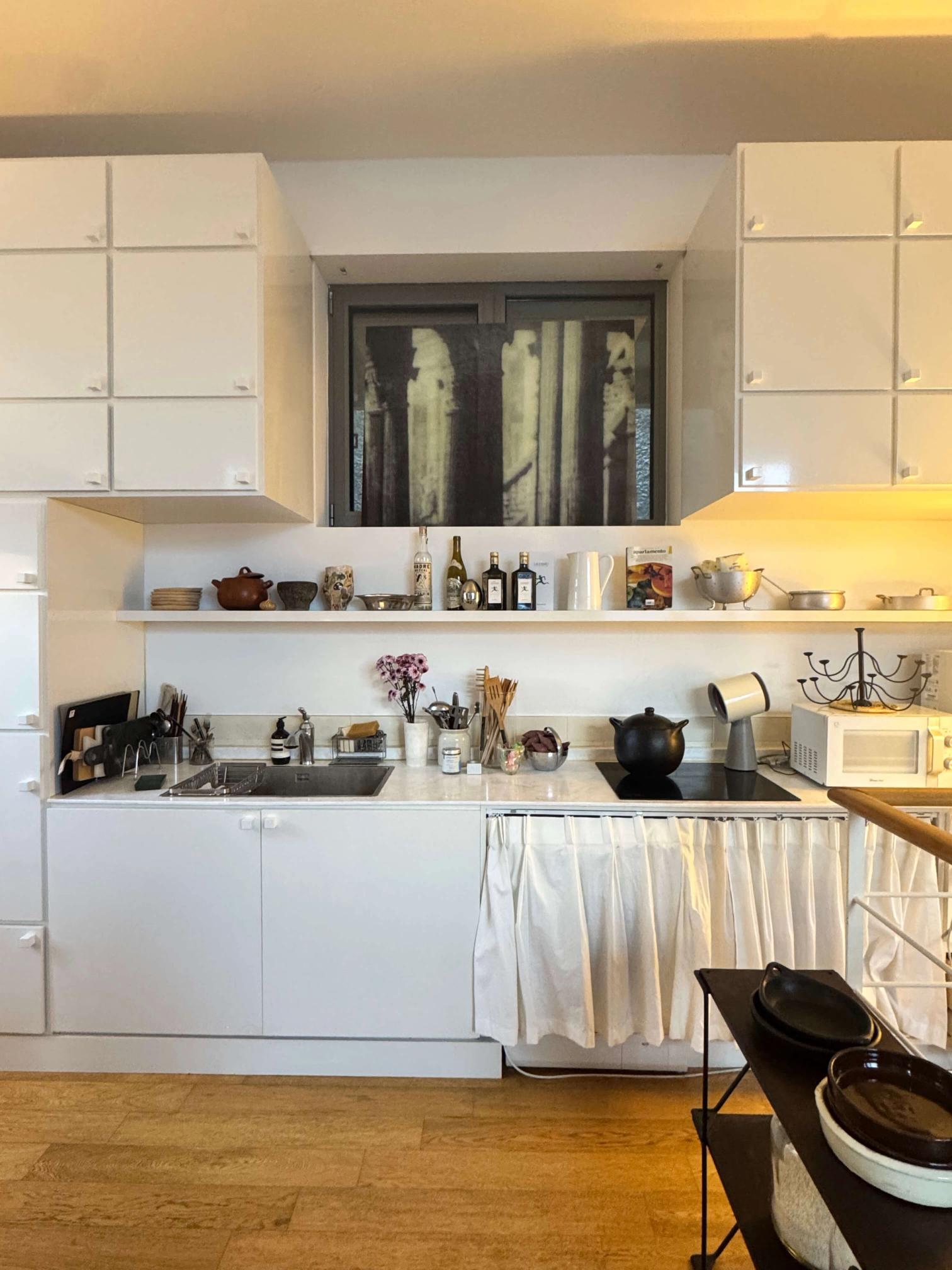 A modern white kitchen with wood floors, an induction cooktop, a sink, and open shelves. A window above the counter features dark abstract art.