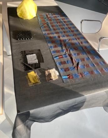 A dark grey table covering displays a yellow crumpled object, a square of black beads, various fabric swatches, and a long piece of blue and reddish-brown striped textile.