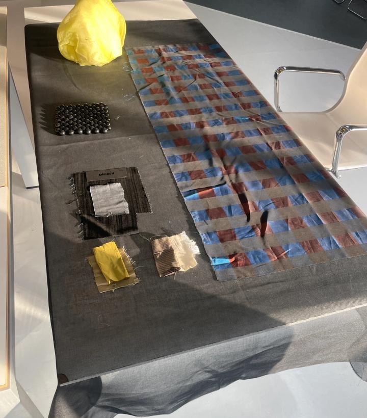 A table draped in dark fabric, holding a crumpled yellow textile, a grid of black beads, assorted fabric swatches, and a long piece of striped sheer fabric.