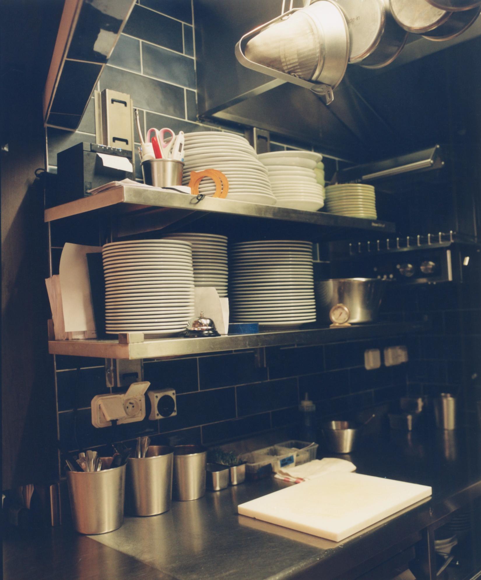 A professional kitchen with black tiled walls, stainless steel counters, and shelves full of stacked plates and tools.