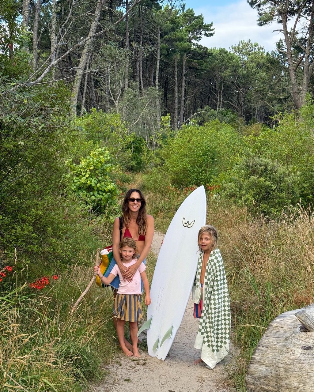 A woman and two children stand on a sandy path with a surfboard, surrounded by lush green foliage and trees.