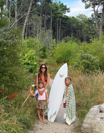 A woman and two children stand on a sandy path with a surfboard, surrounded by lush green foliage and trees.