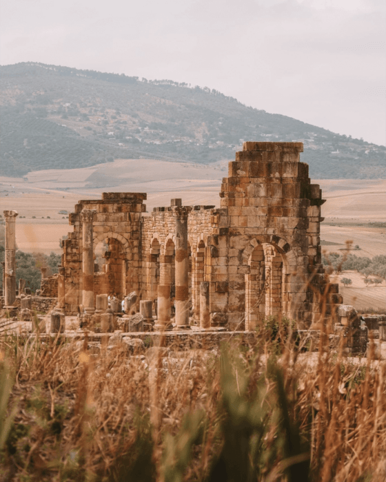 Stone ruins with arches and columns, dry grass in the foreground, and hills and fields in the background.