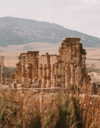 Stone ruins with arches and columns, dry grass in the foreground, and hills and fields in the background.