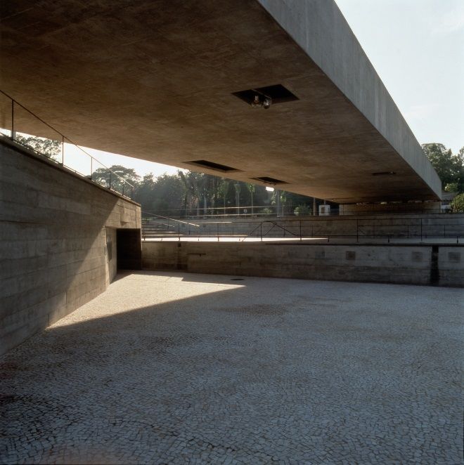 A wide concrete overhang above a large, paved plaza, with strong shadows and concrete walls.