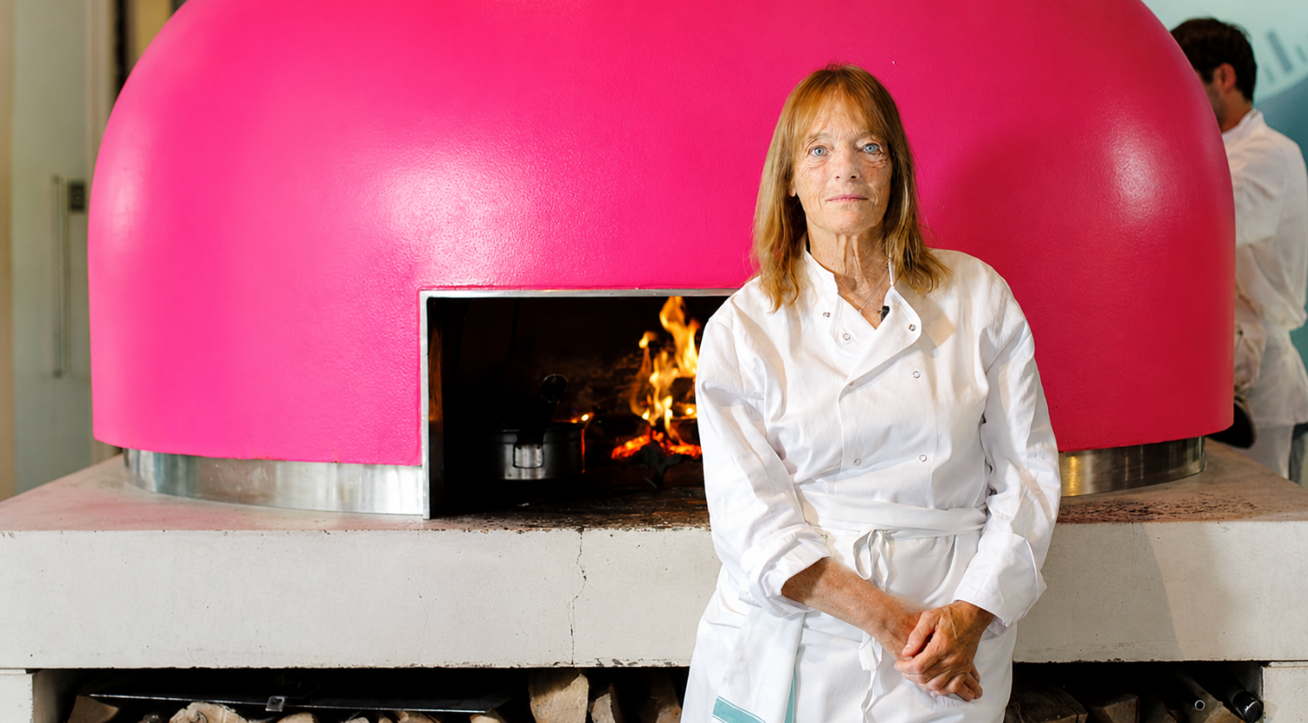 A woman in a chef's uniform stands in front of a bright pink pizza oven with fire burning inside.